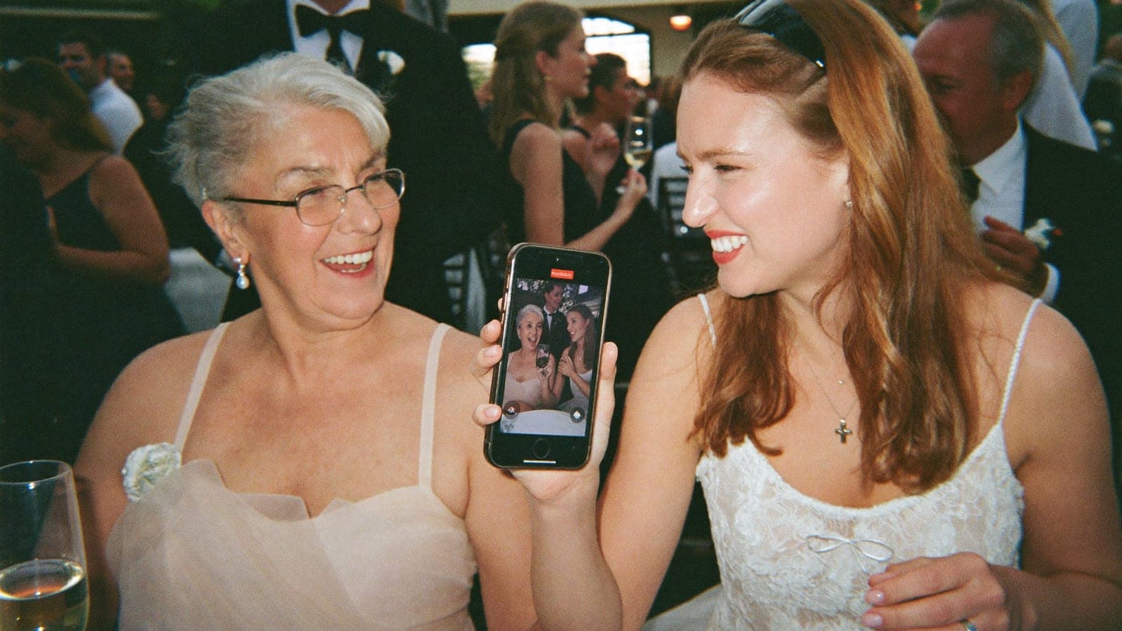 Oma en kleindochter lachen samen naar een telefoon op het bruiloftsfeest