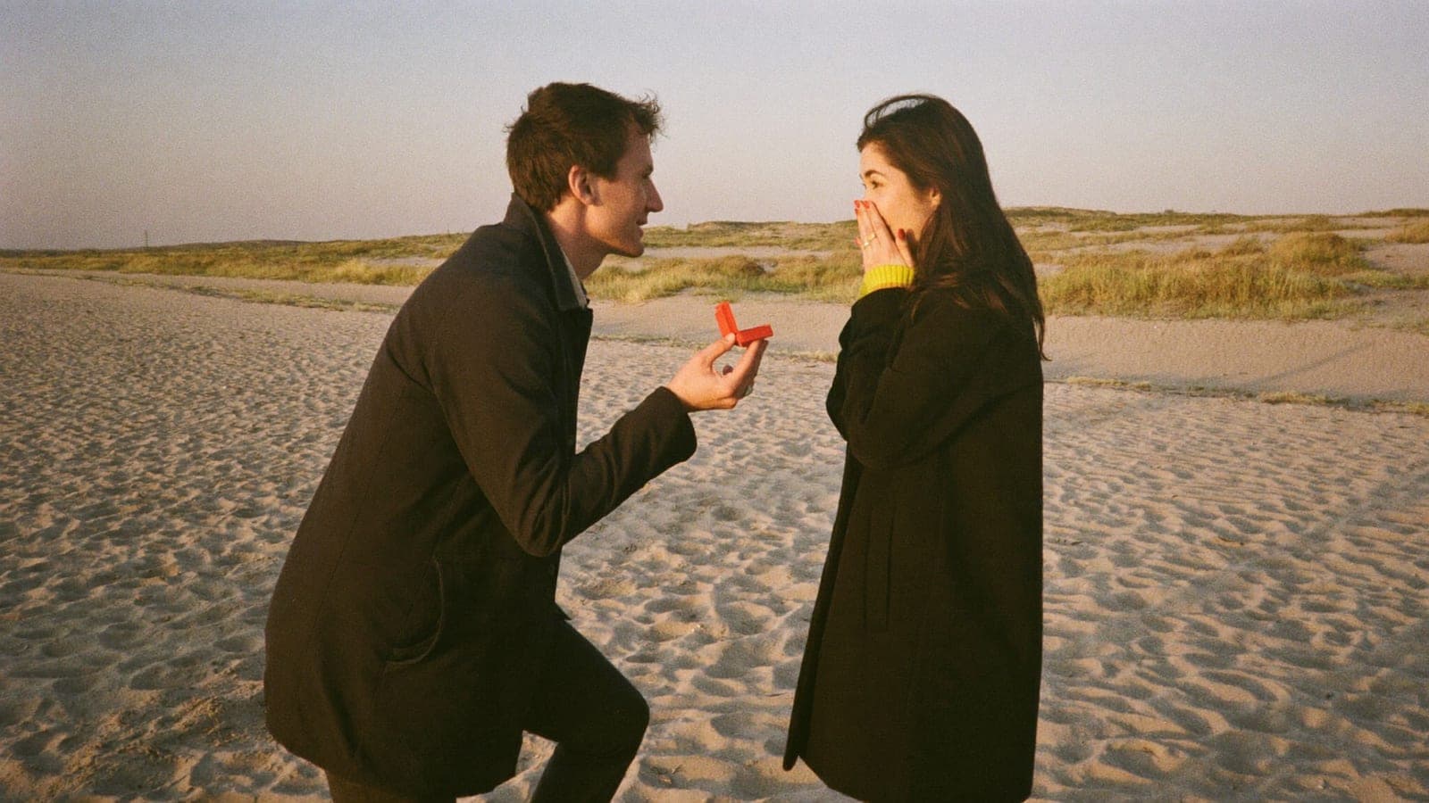 Een man knielt op het strand bij zonsopgang voor een aanzoek, zijn vriendin slaat verrast de hand voor haar mond