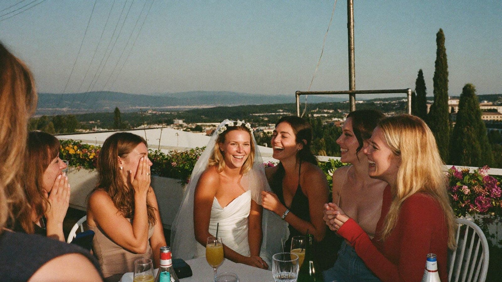 Een groep vriendinnen lacht met cocktails op een dakterras, de bruid met een bloemenkroon